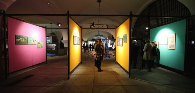 Wystawa w Akademii Sztuk Pięknych w Gdańsku, ekrany ustawione prostopadle do widza od lewej różowy, dwa żółte i zielony, poiedzy nimi zwiedzający; na ekranach wiszą prace plastyczne;